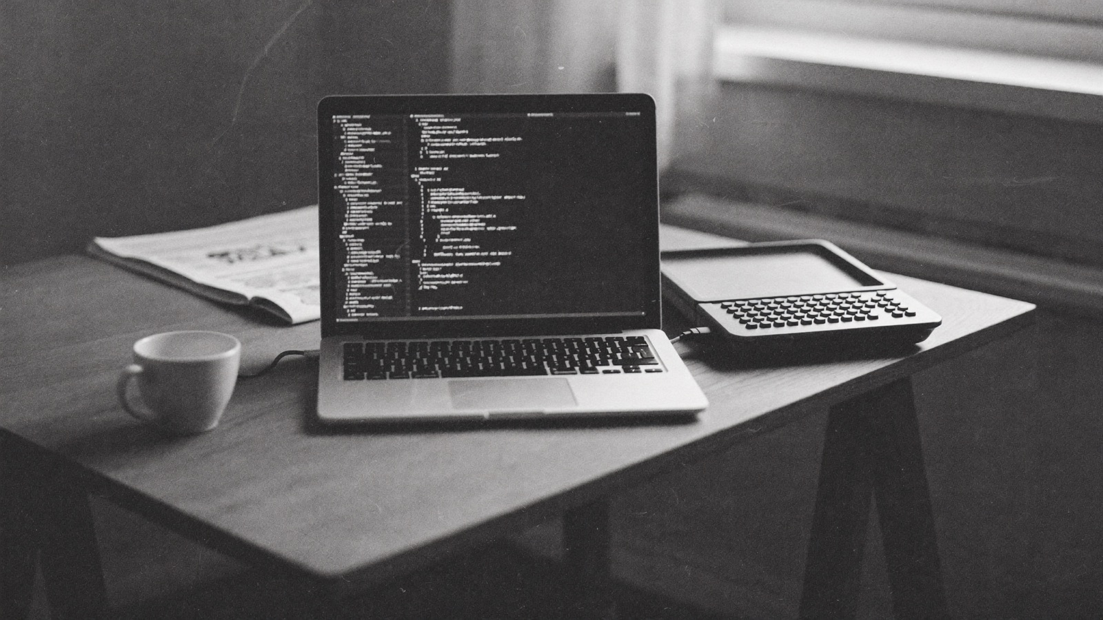 A laptop running a terminal full of code beside a printed newspaper on a wooden desk, morning light